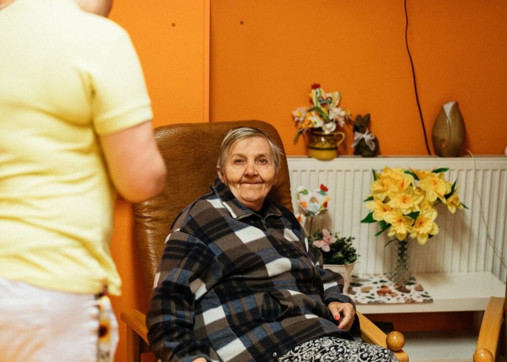 An older resident in an armchair with a caregiver nearby. Illustrative archive, not a CMS enforcement subject.