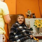 An older resident in an armchair with a caregiver nearby. Illustrative archive, not a CMS enforcement subject.